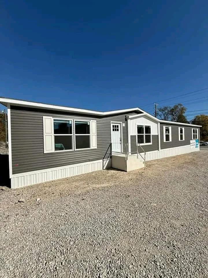 A long, single-story manufactured home with gray siding and white trim, set on a gravel lot under a clear blue sky, conveys a sense of calm and simplicity.