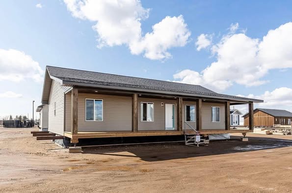 A single-story modular home with a beige exterior and a dark roof sits on a muddy lot under a partly cloudy sky. The house has a wooden porch and multiple windows.