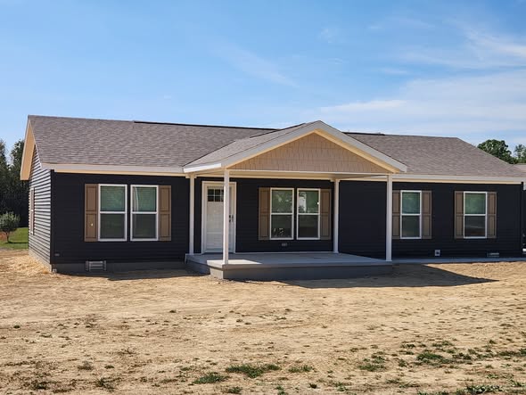 Single-story dark brown house with a gabled roof and white trim. The porch has three columns, surrounded by dry, sandy ground and clear blue sky.