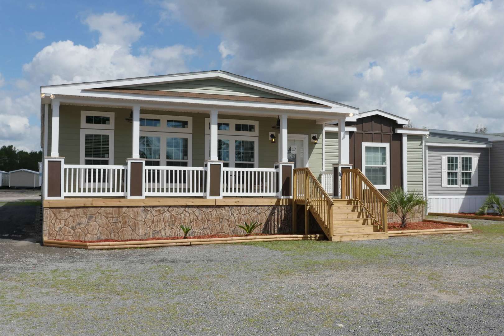 A stylish manufactured home with a stone and white-trimmed facade. It features a wooden porch, steps, and a neatly landscaped area under a cloudy sky.