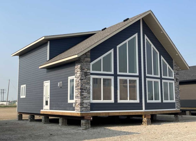 Modern blue house on stilts with large triangular windows and stone accents. It has a peaked roof and a calm, minimalist look on a gravel surface.