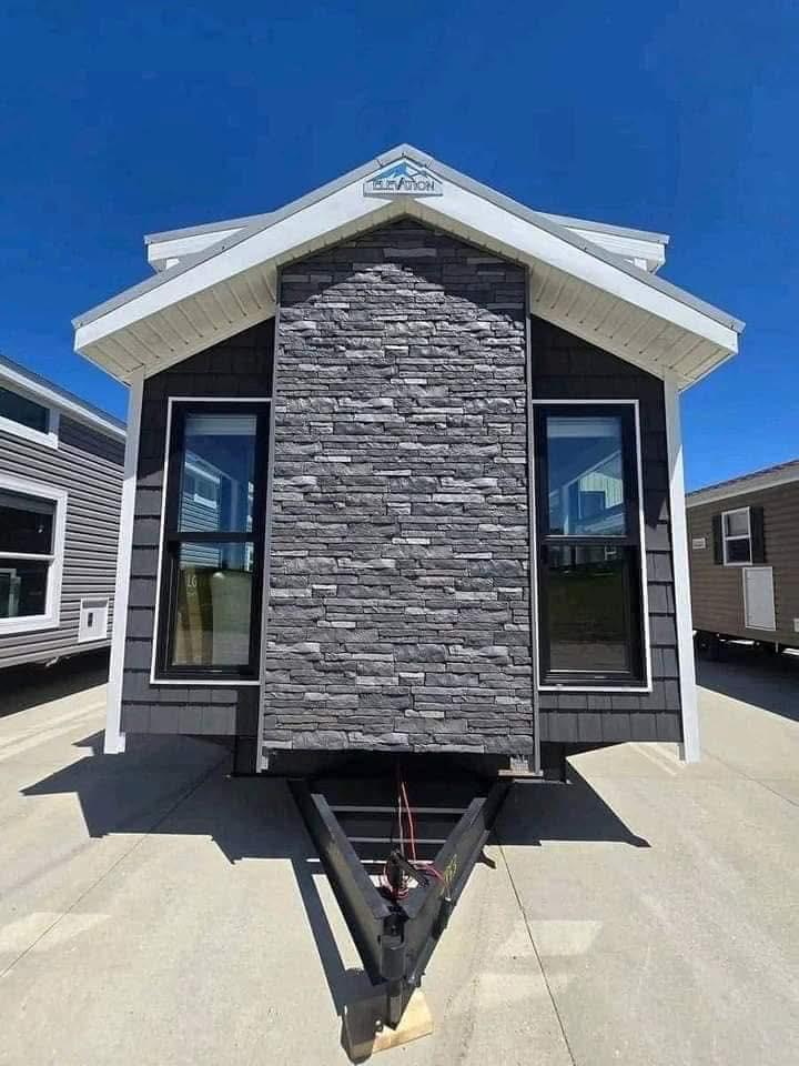 Front view of a modern tiny house on a trailer. Features gray stone facade, white trim, and large windows. Bright blue sky suggests a sunny day.