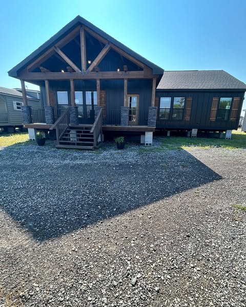A rustic cabin with dark wood and large windows stands under a clear blue sky. Gravel driveway leads to a porch with wooden beams and stairs.