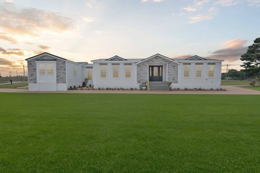 Single-story white house with a stone entrance, surrounded by a lush green lawn at sunset. The sky is partly cloudy, creating a serene ambiance.