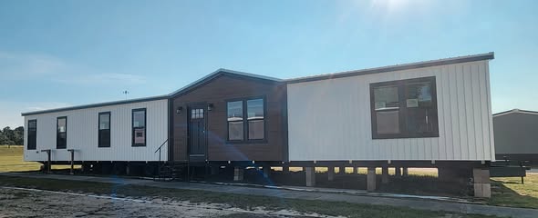 A large, modern mobile home with white and dark wood paneling stands elevated on blocks in a grassy area under clear blue skies.