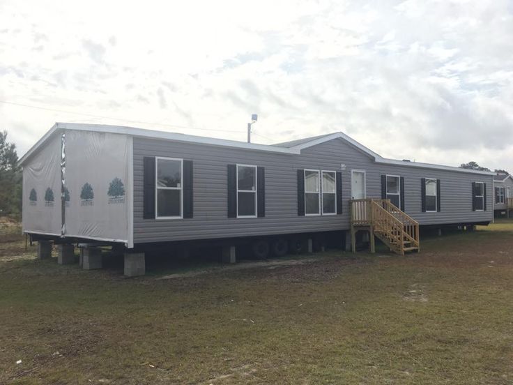 A large, gray manufactured home with white trim and multiple windows on a grassy lot. Wooden stairs lead to the entrance under a cloudy sky.