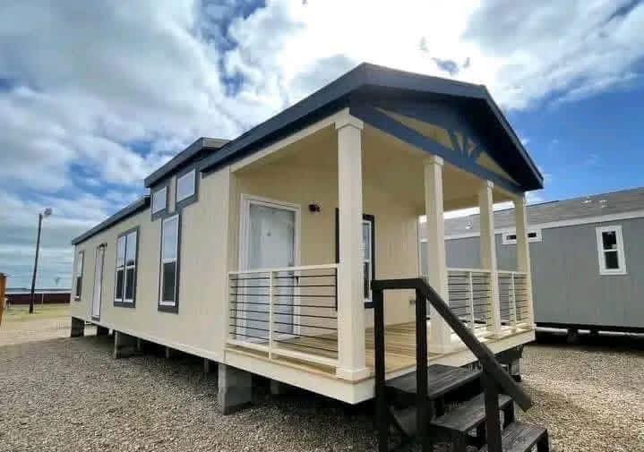 Small beige manufactured home with a porch and dark trim, set on gravel. Bright sky with clouds overhead adds an airy, welcoming feel.