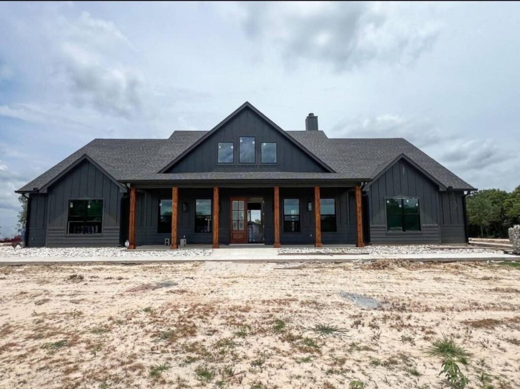 A modern, charcoal grey house with a steep gable roof and wooden columns stands on a sandy plot. The sky is overcast, creating a calm atmosphere.