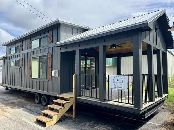 A modern tiny home on wheels with a dark gray exterior, wood accents, large windows, and a small porch with stairs. The scene is bright and inviting.