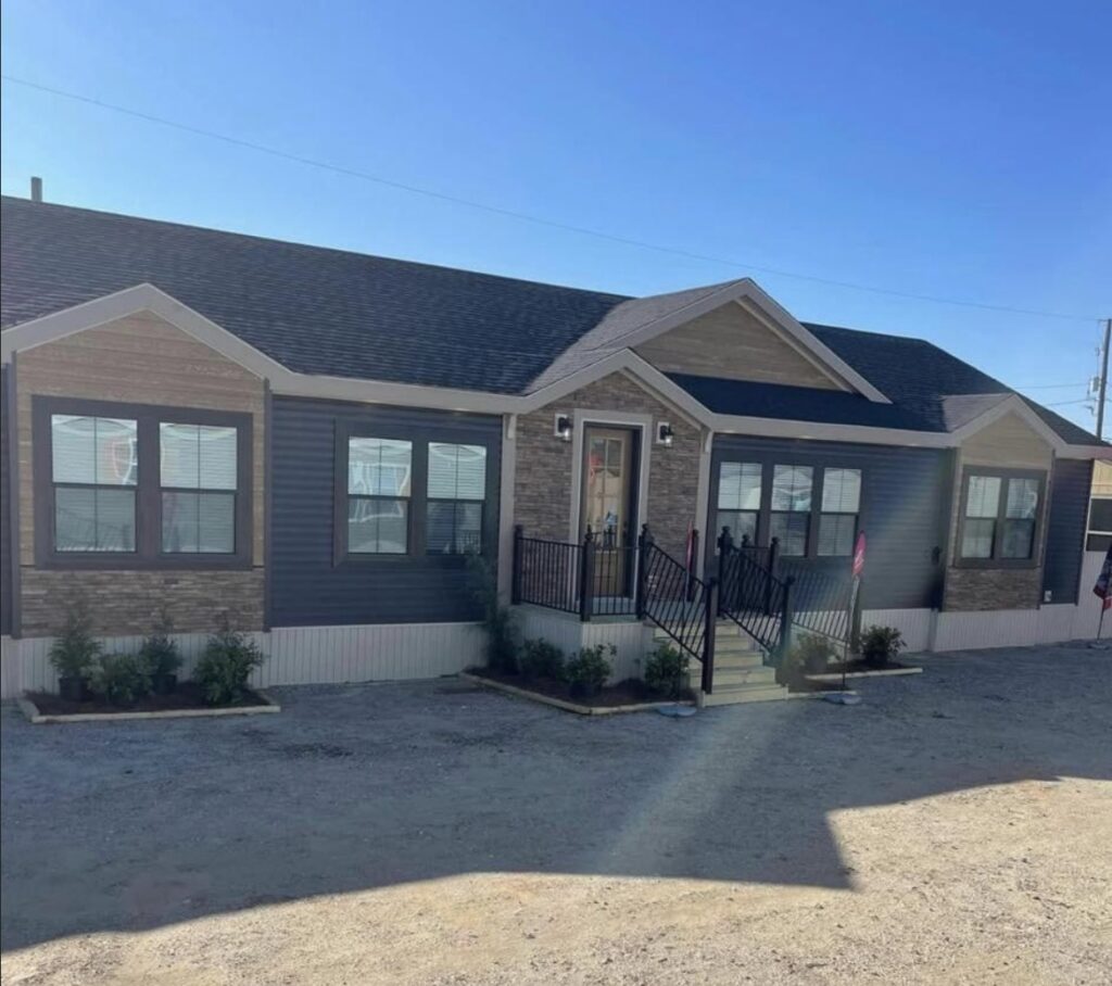 A modern, single-story manufactured home with a mixed stone and dark siding exterior. It features a central porch with stairs and landscaped shrubs. Bright, sunny day.