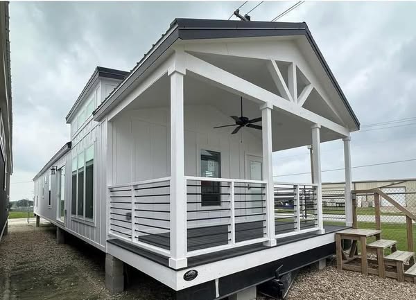 A modern tiny house with a white exterior and gabled roof, featuring a small porch with railing and steps. A ceiling fan is visible, with a cloudy sky.