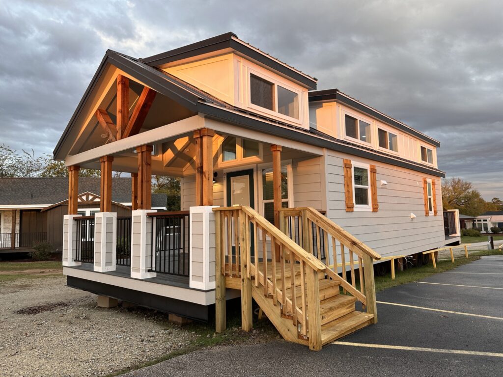 A modern tiny house with a gabled roof and wooden facade is warmly lit at sunset. It has a light gray exterior, wooden accents, and a porch with steps.