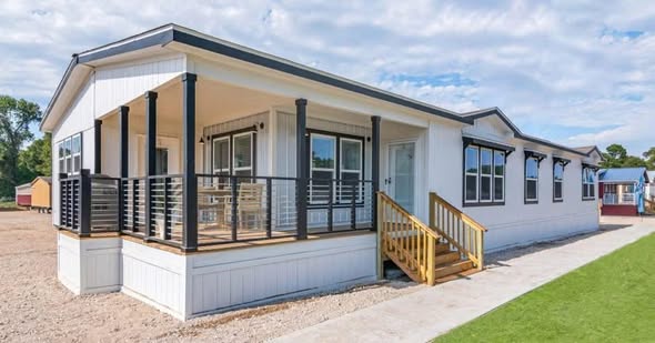 Modern white manufactured home with a black-trimmed porch, wooden steps, and large windows. The setting is sunny with a clear blue sky.