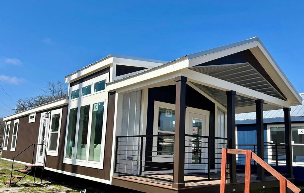 A modern tiny house with a brown and white exterior sits under a blue sky. Large windows and a small porch with metal railings add to its contemporary design.