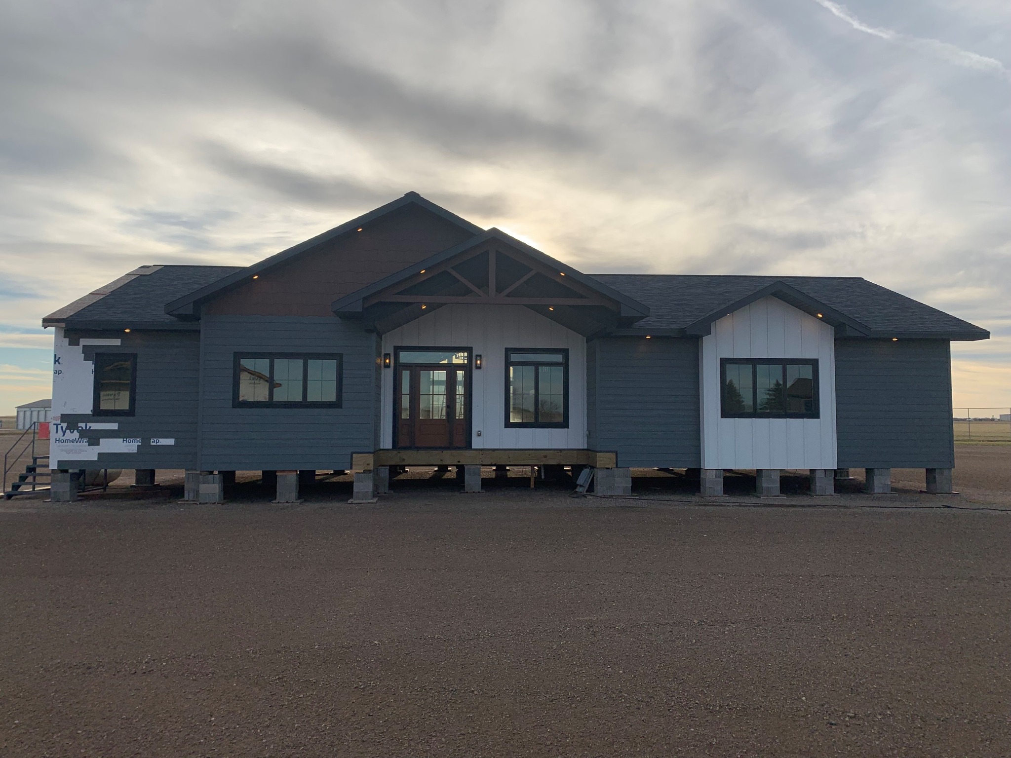Single-story gray and white house with gabled roof stands elevated on stilts against a cloudy sky. A calm, serene tone is conveyed.