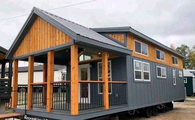 A modern tiny house with a gray exterior and wooden accents. It features a covered porch with wooden pillars and black railings, evoking a cozy, rustic feel.