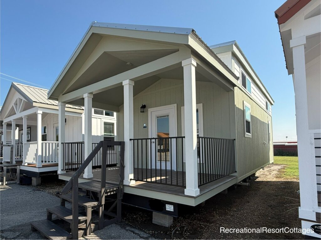 A small, modern light green cottage with a covered porch, white trim, and black railing. It's part of a row of similar homes under a clear blue sky.