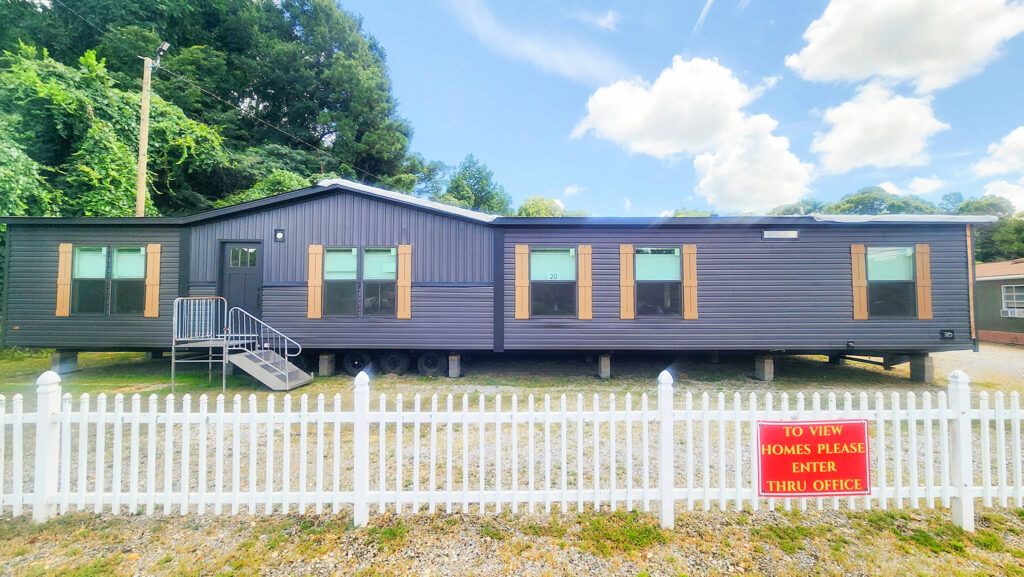 A modern gray mobile home with wooden shutters is surrounded by a white picket fence. A sign on the fence reads, “To view homes please enter thru office.” Lush trees and a bright blue sky with fluffy clouds are visible in the background.