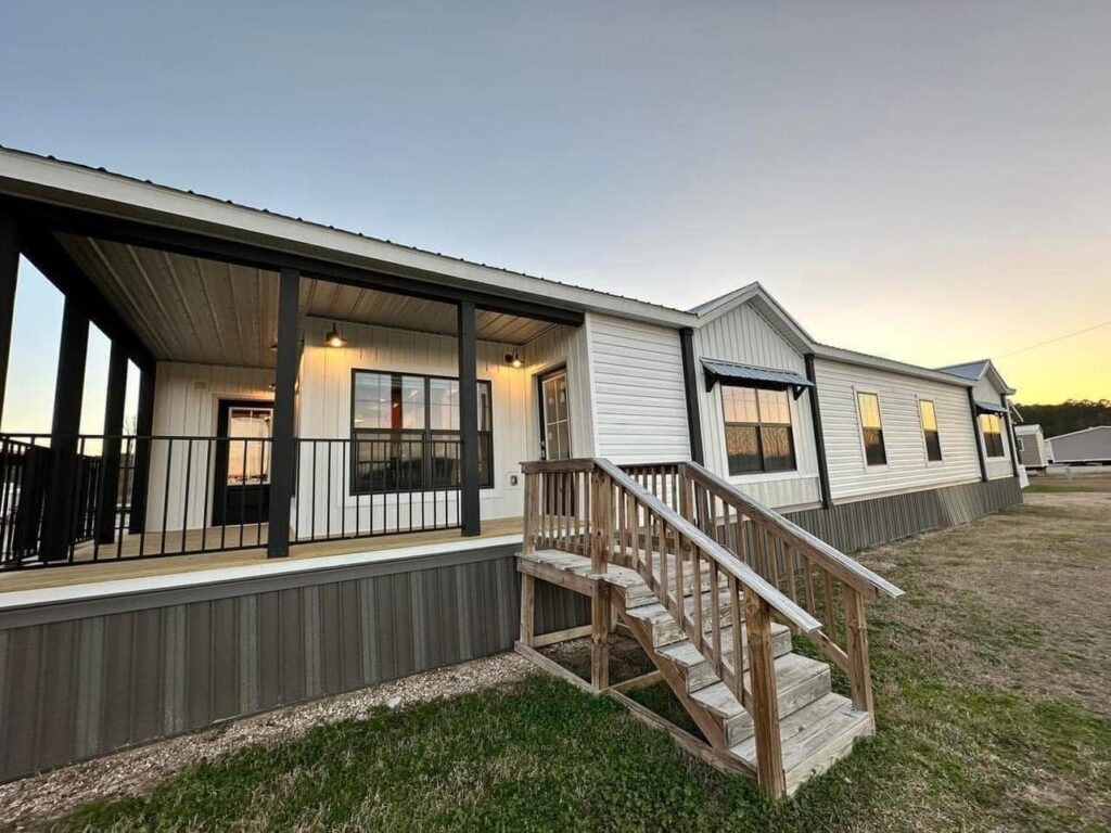 White manufactured home with a wooden porch and stairs, featuring black railings and trim. The setting sun casts a warm glow, creating a cozy atmosphere.