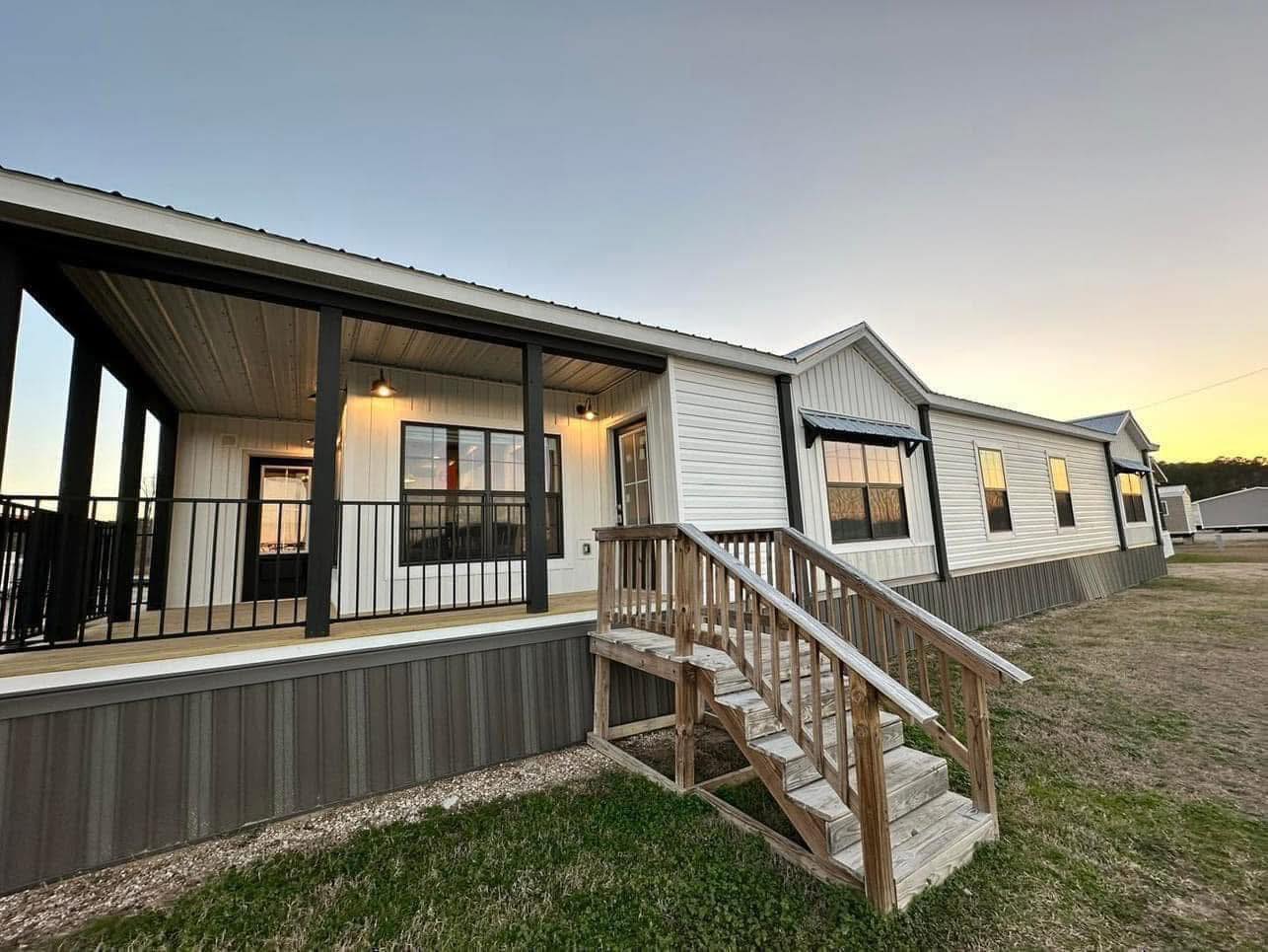White manufactured home with a wooden porch and stairs, featuring black railings and trim. The setting sun casts a warm glow, creating a cozy atmosphere.