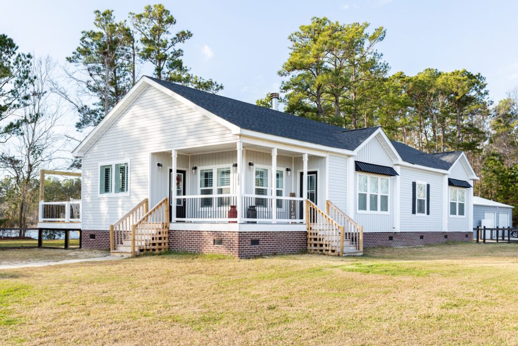 A modern white house with a black roof, set on a grassy lawn surrounded by tall trees. It features a wraparound porch with white railings, creating a welcoming atmosphere.