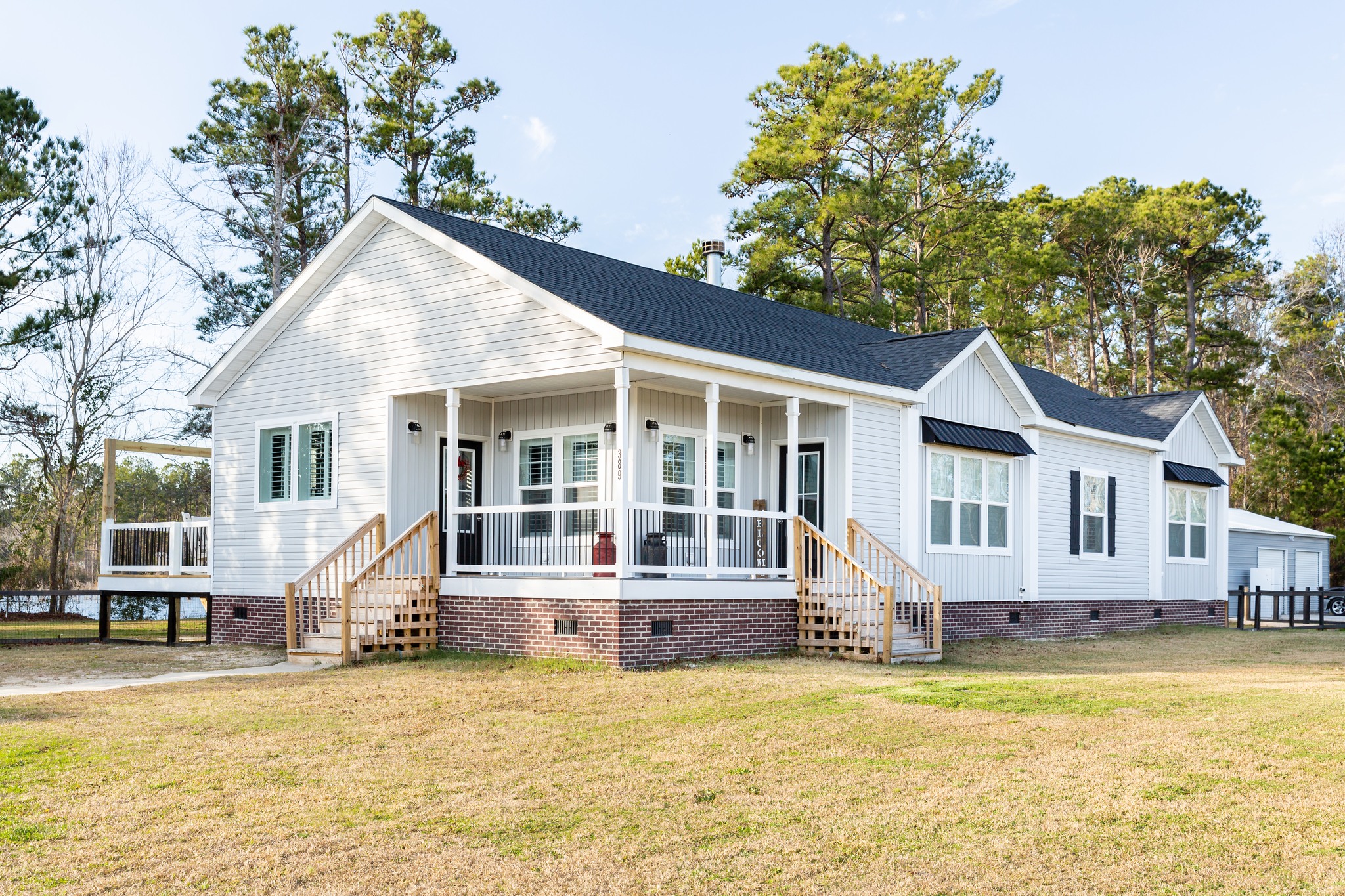 A modern white house with a black roof, set on a grassy lawn surrounded by tall trees. It features a wraparound porch with white railings, creating a welcoming atmosphere.