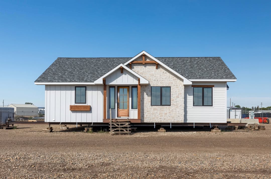 A small, white modern farmhouse with gray roof stands on a gravel lot under a clear blue sky. The facade features wood accents and stone details.