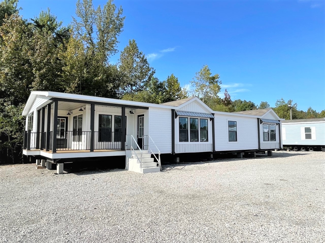 A modern, white modular home sits on a gravel lot, surrounded by trees under a clear blue sky. It features large windows and a small porch with stairs.