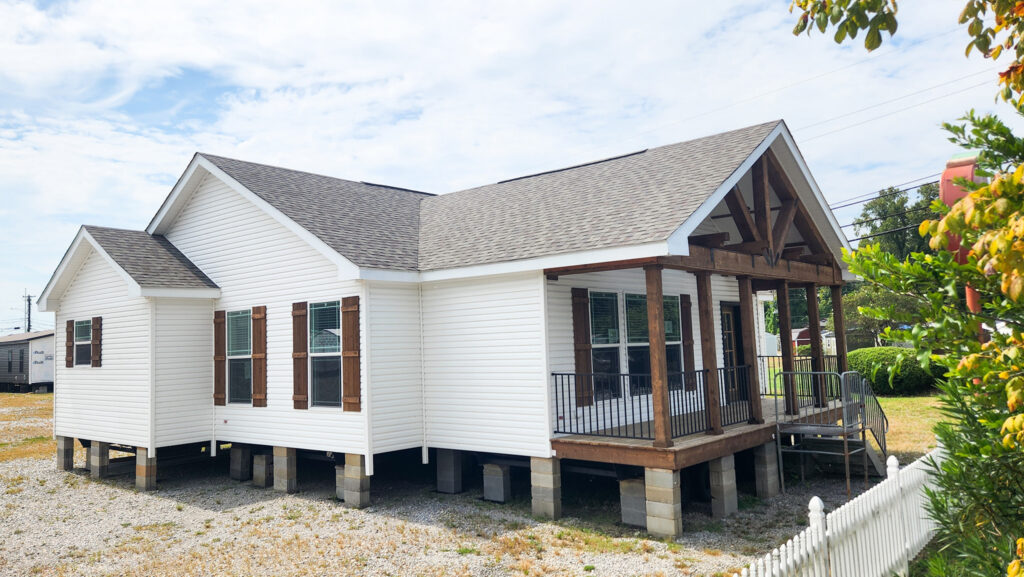 White elevated house with brown shutters and a wooden porch, set against a partly cloudy sky. A white picket fence and greenery are in the foreground.