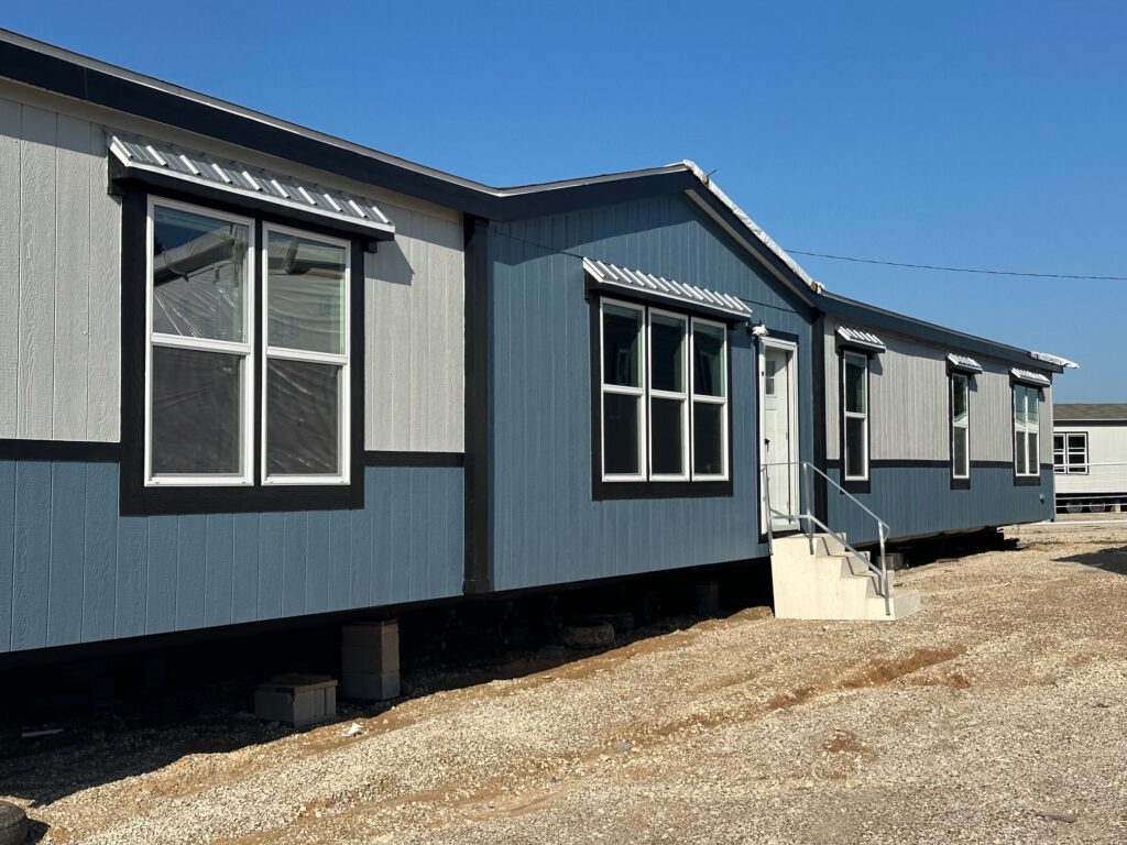 Blue and gray modular building under clear sky, featuring multiple windows and small stairs at the entrance. The setting feels sunny and quiet.