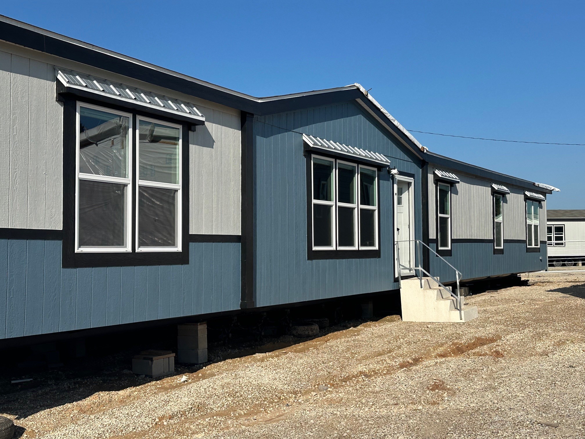 Blue and gray modular building under clear sky, featuring multiple windows and small stairs at the entrance. The setting feels sunny and quiet.