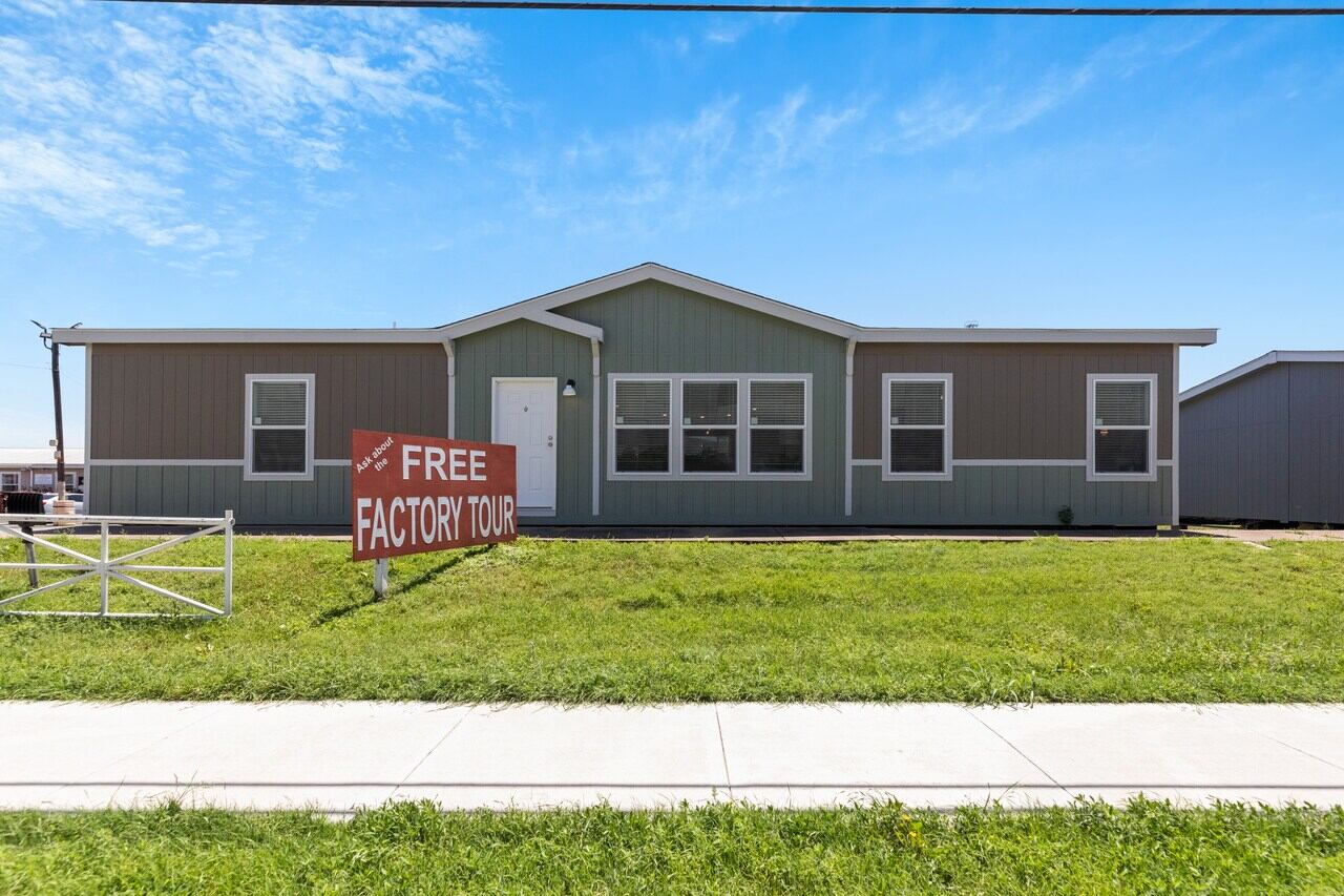 Single-story house with gray siding and six windows, set on a green lawn under a blue sky. A red sign in front reads "Free Factory Tour."