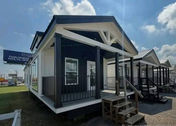 A small, modern white house with a black gabled roof and porch under a blue sky. It has steps leading up, exuding a cozy, welcoming feel.