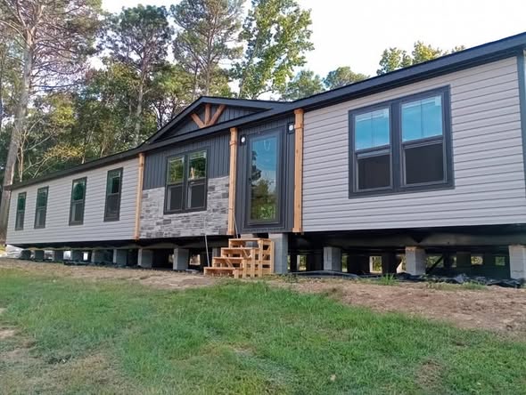 A modern manufactured home with gray siding and stone accents, elevated on blocks. Wooden steps lead to a glass door. Surrounded by grass and trees.