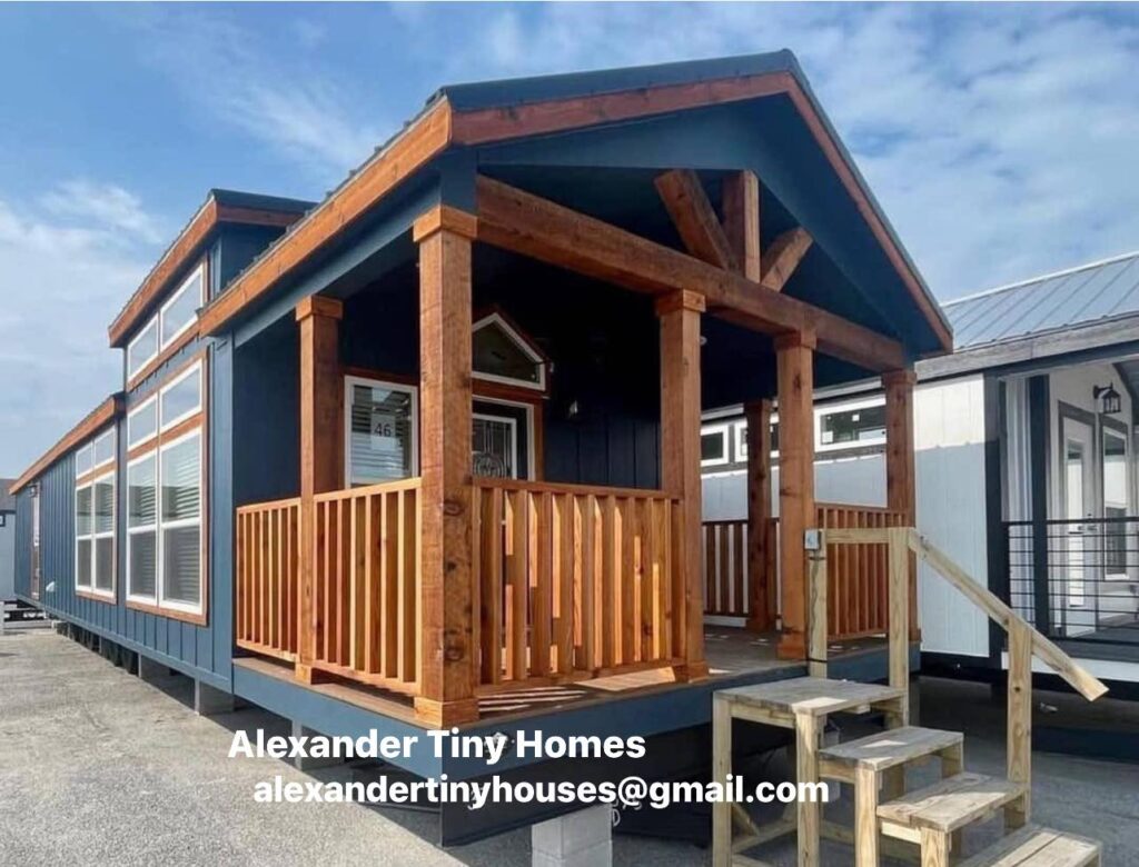 Cozy tiny home with a wooden porch and blue siding under a clear sky. Three steps lead to the entrance. A welcoming, quaint vibe.