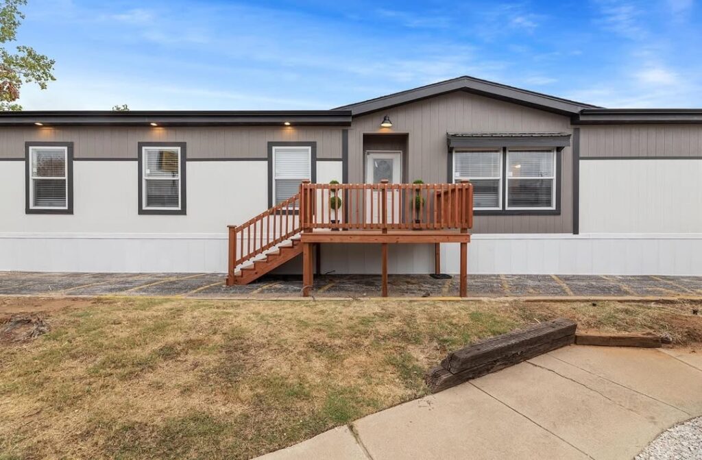 A modern mobile home with a gray and white exterior, featuring a small wooden porch and steps. The sky is clear, conveying a calm, sunny day.