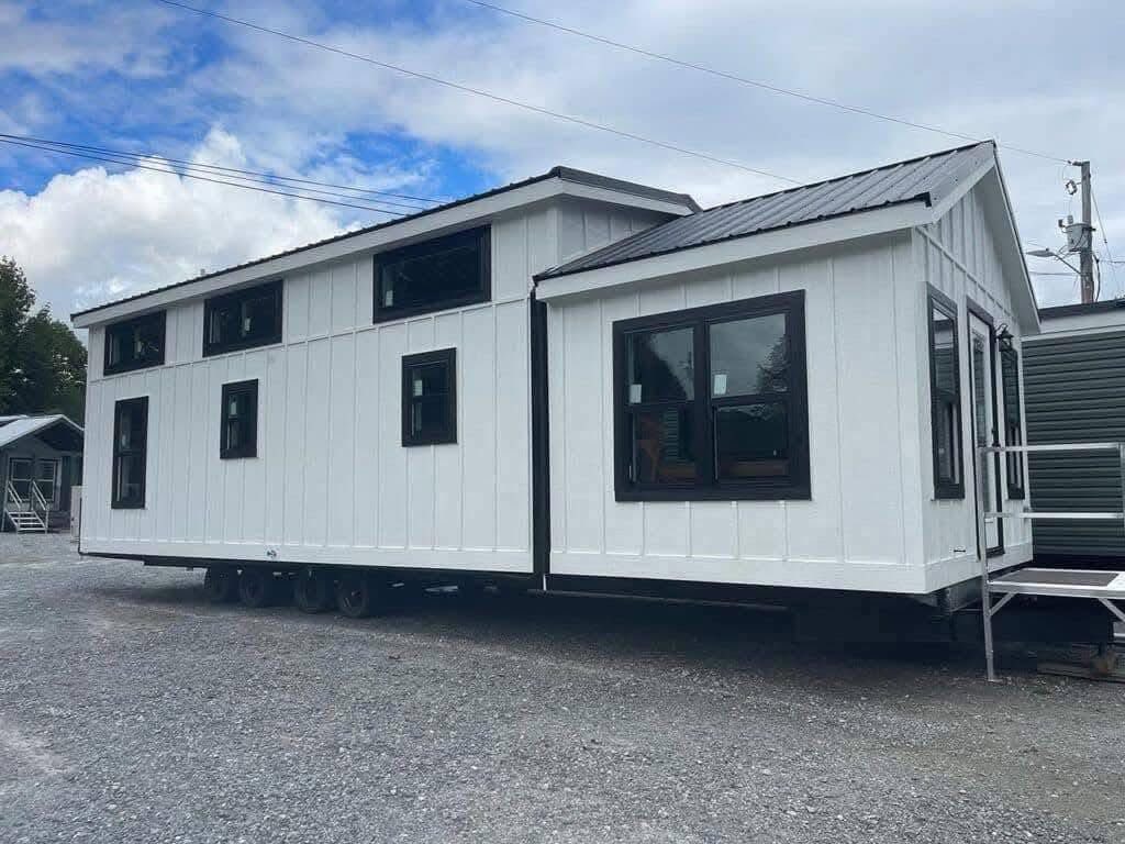 A modern white tiny house on wheels with black trim and windows sits on gravel under a partly cloudy sky, evoking a sense of mobility and minimalism.