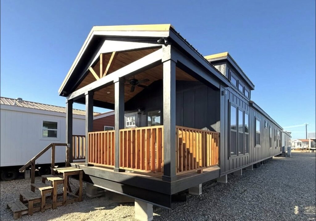 Single-story modern tiny house with a wooden porch and dark exterior, set on a gravel lot. Clear blue sky, conveying a serene and minimalist vibe.