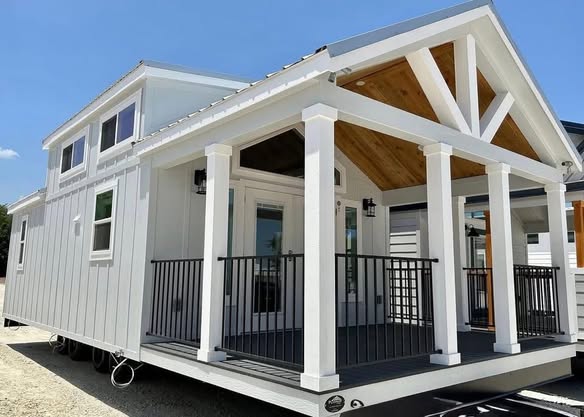 A modern tiny house with white siding and a gabled roof. It features large windows, a covered porch with columns, and clean, minimalistic design elements.