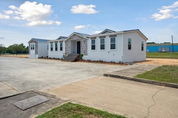 A modern white modular home with multiple sections and gabled roofs sits on a concrete driveway. The sky is clear, giving a calm and spacious feel.