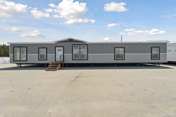 A modern, gray mobile home with large windows and a small set of stairs at the entrance. It's set against a clear blue sky with scattered clouds.
