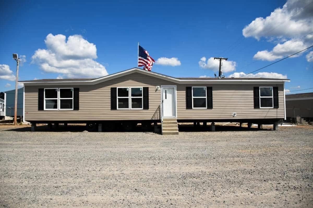 A beige manufactured home with black shutters set on a gravel lot under a blue sky with clouds, featuring an American flag flying on the roof.