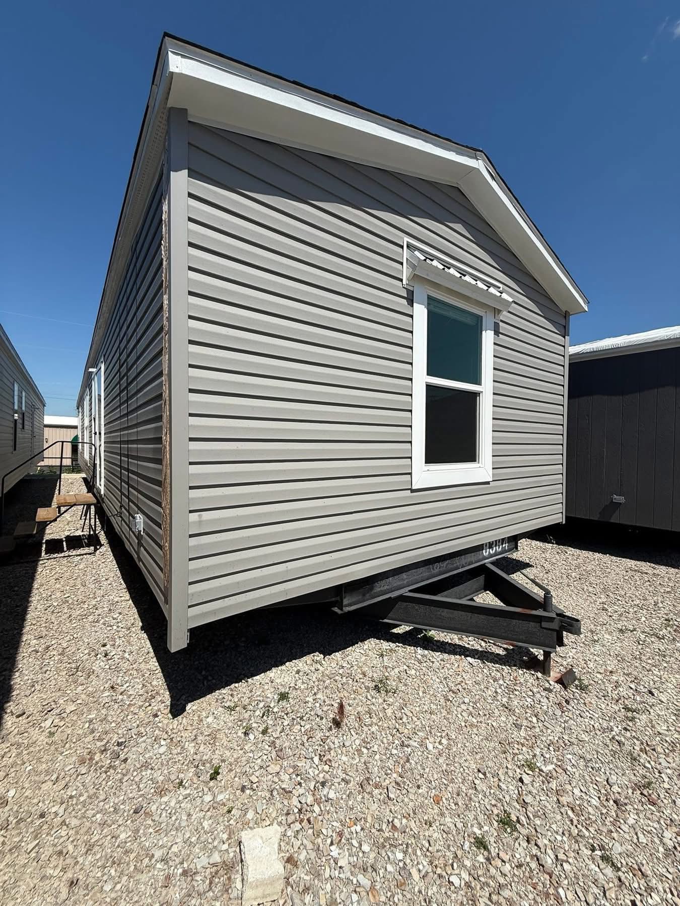 A gray mobile home with white trim and a single window on a gravel lot, under a bright blue sky, conveying a sense of simplicity and mobility.
