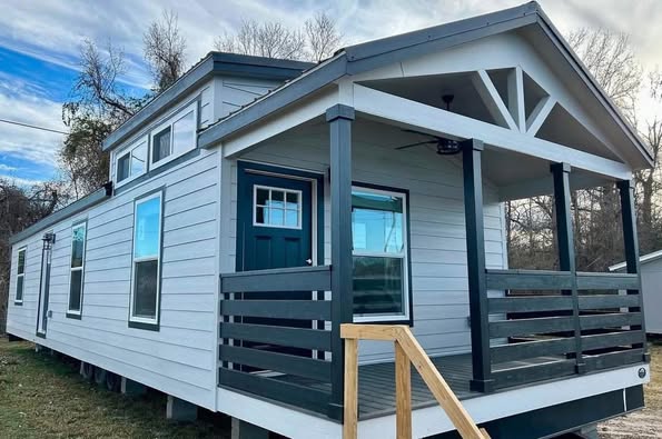 A small, modern tiny house with light gray siding and a white-trimmed blue door on a grassy lot. It has a front porch with dark railings.