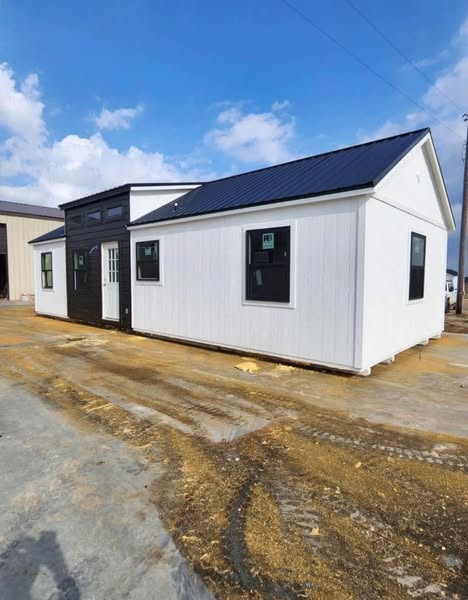 A modern tiny house with a white exterior and black trim stands on a construction site under a blue sky with clouds, conveying a sense of simplicity and new beginnings.
