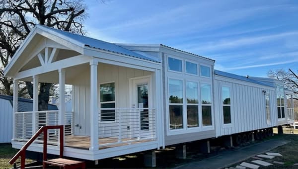 A modern white modular home with large windows and a metal roof, set on a raised platform. The entrance features a small porch with a railing and red steps, under a clear blue sky.