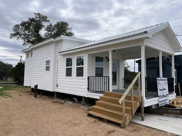 White tiny house with a gabled roof and covered porch, situated on a gravel lot. The overcast sky and distant trees create a calm, simple atmosphere.