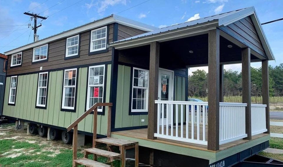 A green and brown tiny house on wheels with a porch featuring white railings and wooden steps, set on grass against a clear blue sky backdrop.