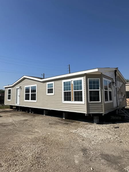 A beige mobile home with multiple windows sits elevated above a gravel lot under a clear blue sky, evoking a sense of simplicity and tranquility.