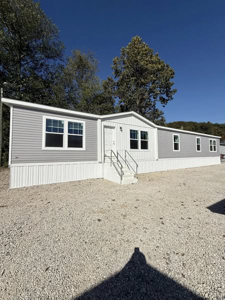 A light gray mobile home with white trim and a central white door, set on a gravel area. Trees and a clear blue sky are in the background.